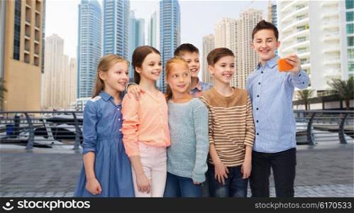 childhood, friendship, technology and people concept - group of happy children talking selfie by smartphone over dubai city street background
