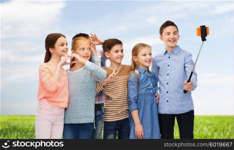 childhood, friendship, technology and people concept - happy children talking picture by smartphone on selfie stick over blue sky and grass background