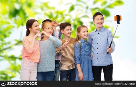 childhood, friendship, technology and people concept - happy children talking picture by smartphone on selfie stick over green natural background