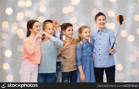 childhood, friendship, technology and people concept - happy children talking picture by smartphone on selfie stick over holidays lights background