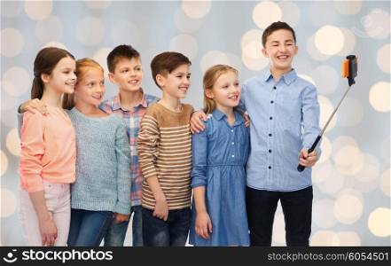 childhood, friendship, technology and people concept - happy children talking picture by smartphone on selfie stick over holidays lights background