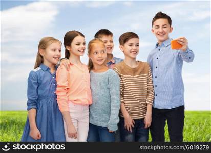 childhood, friendship, technology and people concept - happy children talking selfie by smartphone over blue sky and grass background