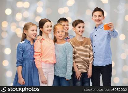 childhood, friendship, technology and people concept - happy children talking selfie by smartphone over holidays lights background