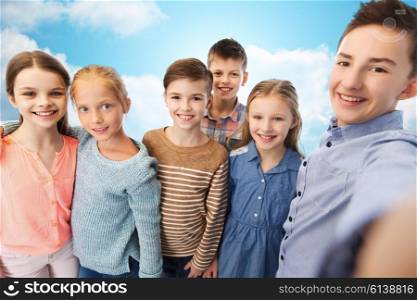 childhood, friendship, technology and people concept - happy children talking selfie over blue sky and clouds background