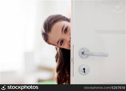 childhood, fun and people concept - happy smiling beautiful girl hiding behind door at home. happy smiling beautiful girl behind door at home