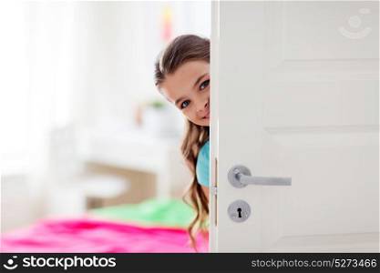 childhood, fun and people concept - happy smiling beautiful girl hiding behind door at home. happy smiling beautiful girl behind door at home