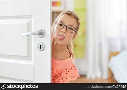 childhood, fun and people concept - happy smiling beautiful girl in glasses behind door sticking out her tongue at home. girl behind door showing her tongue at home