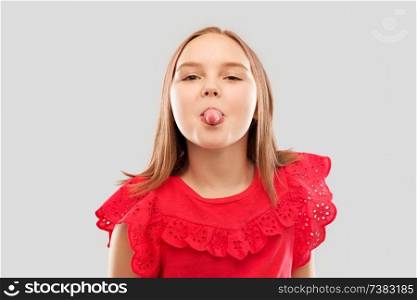 childhood, gesture and people concept - beautiful smiling girl in red shirt showing her tongue over grey background. beautiful girl in red shirt showing her tongue