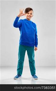 childhood, gesture and people concept - portrait of smiling boy in blue hoodie showing ok hand sign over grey background. boy in blue hoodie showing ok hand sign