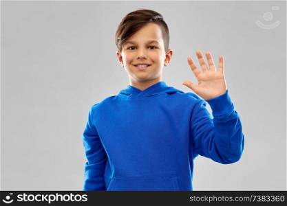 childhood, gesture and people concept - portrait of smiling boy in blue hoodie waving hand over grey background. smiling boy in blue hoodie waving hand