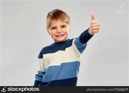 childhood, gesture and people concept - portrait of smiling little boy in striped pullover showing thumbs up over grey background. little boy in striped pullover showing thumbs up