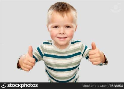 childhood, gesture and people concept - portrait of smiling little boy in striped shirt showing thumbs up over grey background. smiling boy in striped shirt showing thumbs up