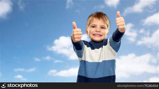 childhood, gesture and people concept - portrait of smiling little boy in striped pullover showing thumbs up over blue sky and clouds background. little boy in striped pullover showing thumbs up