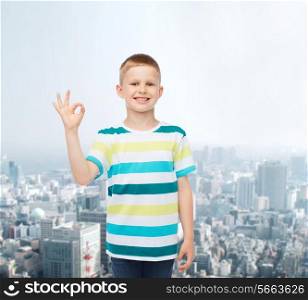 childhood, gesture and people concept - smiling little boy in casual clothes making ok gesture over city background