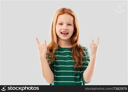 childhood, gesture and people concept - smiling red haired girl in green striped shirt showing rock hand sign over grey background. smiling red haired girl showing rock gesture
