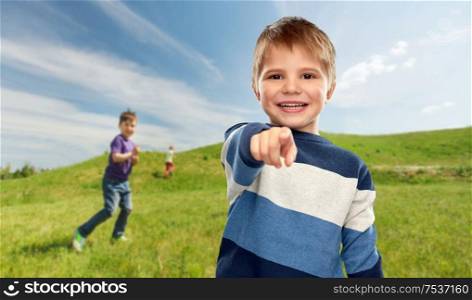 childhood, gesture and summer concept - portrait of smiling little boy in striped pullover pointing finger over blue sky and meadow background. little boy pointing finger to camera outdoors