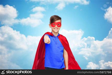 childhood, gesture, carnival costume and people concept - happy boy in red superhero cape and mask pointing finger to you over blue sky and clouds background