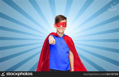 childhood, gesture, carnival costume and people concept - happy boy in red superhero cape and mask pointing finger to you over blue burst rays background