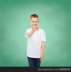 childhood, gesture, education and advertisement concept - smiling boy in white t-shirt pointing finger at you over green board background