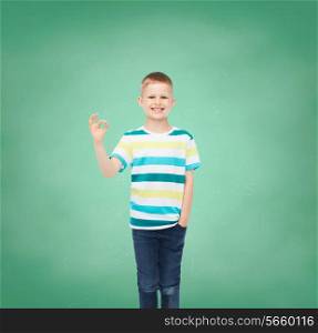 childhood, gesture, education and people concept - smiling little boy in casual clothes making OK gesture over green board background