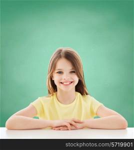 childhood, happiness, education, school and people concept - beautiful little girl sitting at table over green blackboard background
