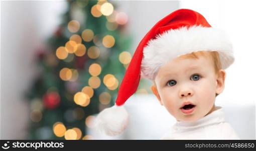 childhood, holidays and people concept - beautiful little baby boy in christmas santa hat over christmas tree lights background