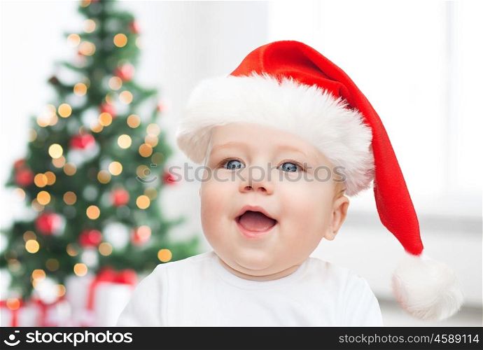 childhood, holidays and people concept - beautiful little baby boy in christmas santa hat over living room with christmas thee lights background