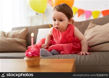 childhood, holidays and people concept - happy baby girl blowing to candle on cupcake at home birthday party. girl blowing to candle on birthday cupcake at home
