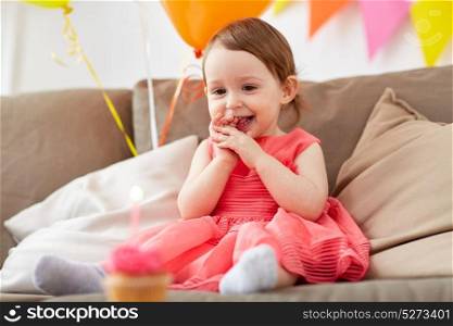 childhood, holidays and people concept - happy baby girl looking at candle on cupcake at home birthday party. happy baby girl looking at birthday cupcake