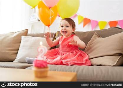 childhood, holidays and people concept - happy baby girl on birthday party at home with air balloons, garland and cupcake with candle. happy baby girl on birthday party at home