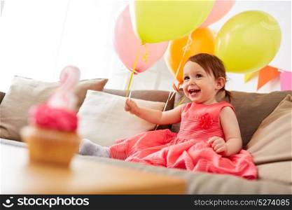 childhood, holidays and people concept - happy baby girl with air balloons, garland and cupcake on birthday party at home. happy baby girl on birthday party at home