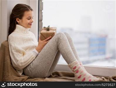 childhood, holidays and people concept - happy beautiful girl with christmas gift sitting on sill at home window. girl with christmas gift sitting on sill at home