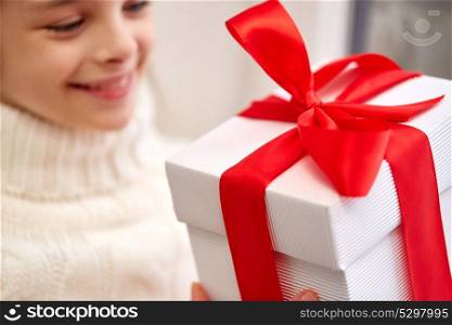 childhood, holidays and people concept - happy beautiful girl with christmas gift sitting on sill at home window. girl with christmas gift sitting on sill at home