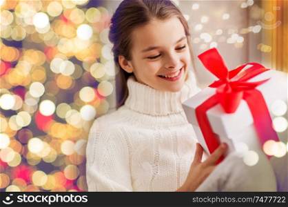 childhood, holidays and people concept - happy beautiful girl with christmas gift box over festive lights background. happy beautiful girl with christmas gift at home