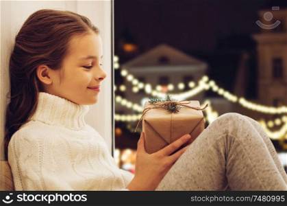 childhood, holidays and people concept - happy beautiful girl with christmas gift sitting on sill at home window over festive street lights background. girl with christmas gift sitting at window