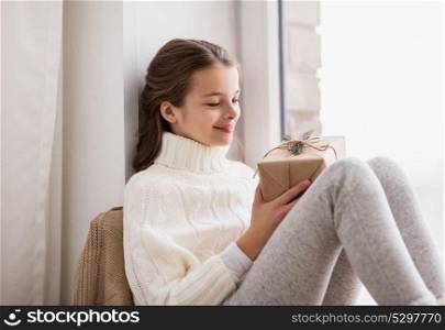 childhood, holidays and people concept - happy beautiful girl with christmas gift sitting at home window. girl with christmas gift sitting at home