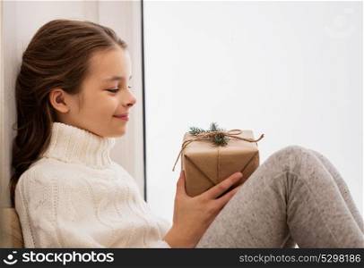 childhood, holidays and people concept - happy beautiful girl with christmas gift sitting at home window. girl with christmas gift sitting at home