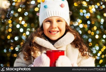 childhood, holidays and season concept - happy little girl with cup of hot tea over christmas tree lights background. little girl with cup of hot tea in winter park