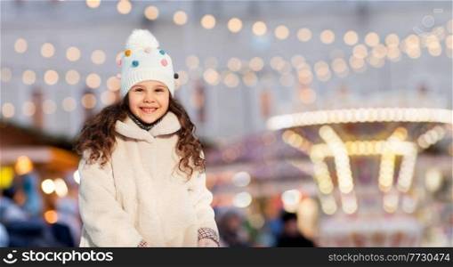 childhood, holidays and season concept - portrait of happy little girl in winter clothes outdoors at park over christmas market or amusement park lights background. happy little girl over christmas market lights