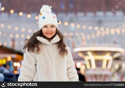 childhood, holidays and season concept - portrait of happy little girl in winter clothes outdoors at park over christmas market or amusement park lights background. happy little girl over christmas market lights
