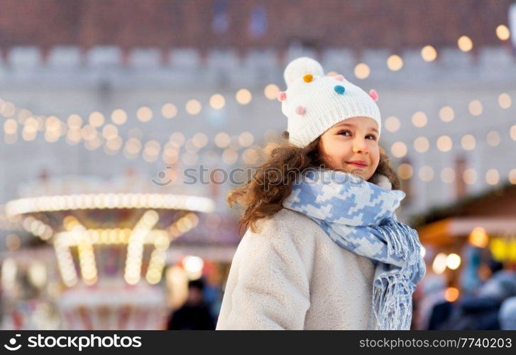 childhood, holidays and season concept - portrait of happy little girl in winter clothes outdoors at park over christmas market or amusement park lights background. happy little girl over christmas market lights