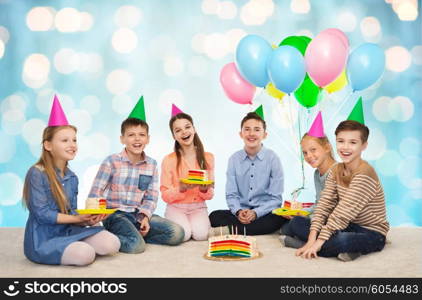 childhood, holidays, celebration, friendship and people concept - happy smiling children in party hats with birthday cake and balloons over blue holidays lights background
