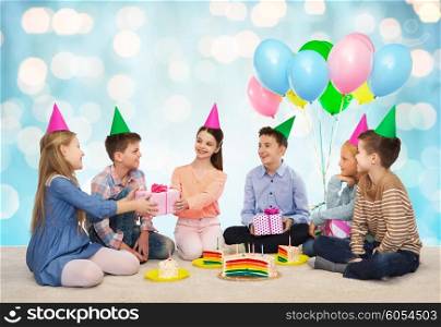 childhood, holidays, celebration, friendship and people concept - happy smiling children in party hats with cake giving presents at birthday party over blue holidays lights background