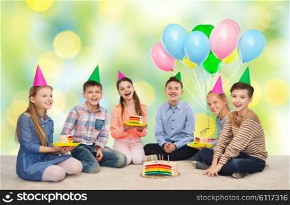 childhood, holidays, celebration, friendship and people concept - happy smiling children in party hats with birthday cake and balloons over green summer holidays lights background
