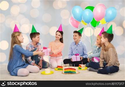 childhood, holidays, celebration, friendship and people concept - happy smiling children in party hats with cake giving presents at birthday party over lights background