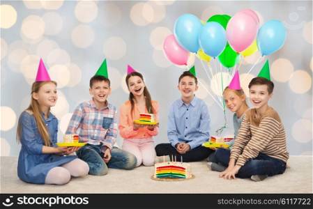childhood, holidays, celebration, friendship and people concept - happy smiling children in party hats with birthday cake and balloons over lights background