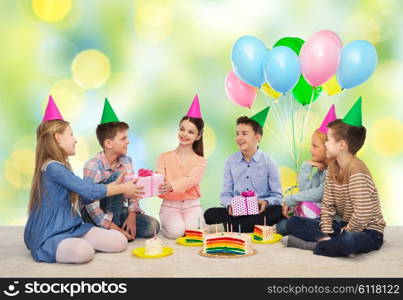 childhood, holidays, celebration, friendship and people concept - happy smiling children in party hats with cake giving presents at birthday party over green summer holidays lights background