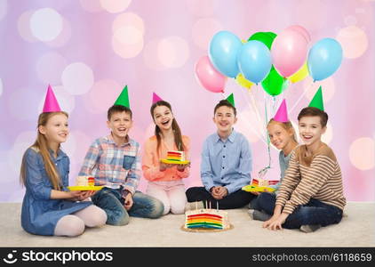 childhood, holidays, celebration, friendship and people concept - happy smiling children in party hats with birthday cake and balloons over pink holidays lights background