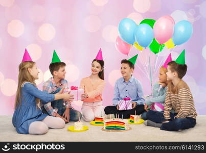 childhood, holidays, celebration, friendship and people concept - happy smiling children in party hats with cake giving presents at birthday party over pink holidays lights background