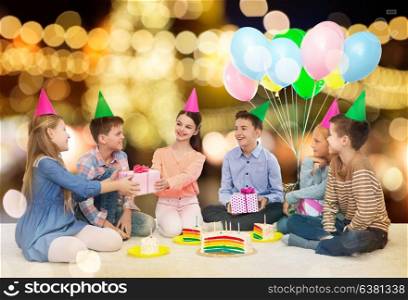 childhood, holidays, celebration, friendship and people concept - happy smiling children in party hats with cake giving presents at birthday party over festive lights background. happy children giving presents at birthday party
